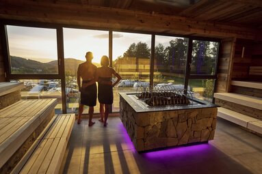 Eine Frau und ein Mann stehen Arm in Arm an der Glasfront der Sauna und blicken auf den Sonnenuntergang. | © Bodenmais Tourismus & Marketing GmbH