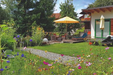 Über eine Blumenwiese hinweg blickt man in einen schön gestalteten Garten. auf der einen Seite steht eine Hängeliege. Auf der anderen Seite befinden sich noch weitere Liegen, teilweise auf einer Terrasse mit Schirm. | © Bodenmais Tourismus & Marketing GmbH