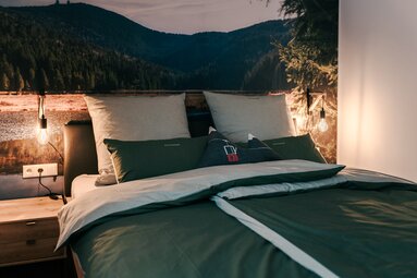 An einer Wand mit einem Bild vom Großen Arber steht ein Holzbett mit grüner Bettwäsche. | © Bodenmais Tourismus & Marketing GmbH