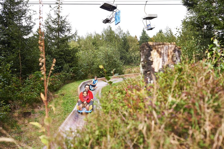 Ein Vater sitzt mit seinem Kind in einem Bob auf einer Sommerrodelbahn. Die beiden fahrend lachend den Berg hinunter. Über der Rodelbahn fährt der Sessellift. | © Bodenmais Tourismus & Marketing GmbH