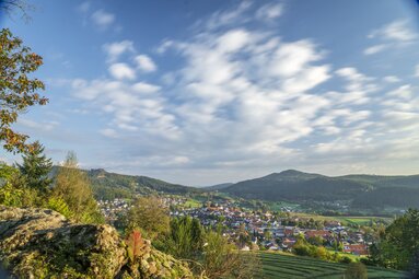 Hinter Bäumen und Hügeln versteckt liegt Bodenmais. Vor der Ortschaft ist eine gemähte Wiese zu erkennen. Am blauen Himmel zeichnen sich weißen Wolken ab. | © Bodenmais Tourismus & Marketing GmbH
