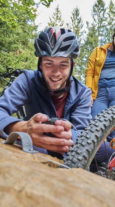 Eine Frau und ein Mann haben ihre Fahrräder an einem Fahrradständer aus Holz abgestellt. Der Mann schließt sein Fahrrad ab und fädelt die Kette durch eine Metallöse am Baumstamm. | © Bodenmais Tourismus & Marketing GmbH