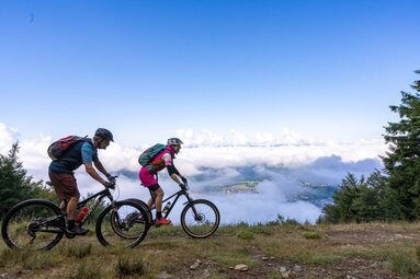 Eine Frau und ein Mann fahren mit dem Mountainbike einen Weg entlang. Hinter ihnen sind prächtige weiße Wolken zu sehen. Dazwischen  kann man auf Bodenmais hinab blicken. | © Bodenmais Tourismus & Marketing GmbH