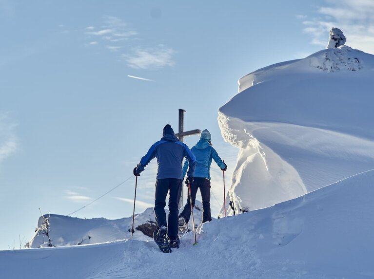 Zwei Wanderer mit Schneeschuhen und Stöcken wandern auf ein Gipfelkreuz zu. Alles ist mit Schnee bedeckt und rechts vom Weg haben sich große Schneeformationen gebildet. | © Bodenmais Tourismus & Marketing GmbH