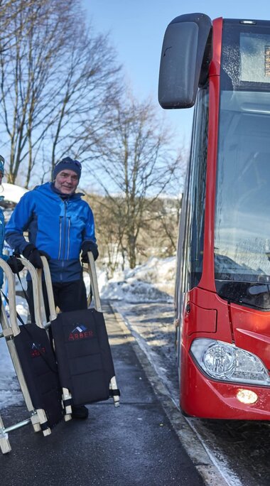 Ein roter Bus mit der Anzeige "Bretterschachten" fährt gerade in die Haltestelle ein. Dort stehen ein Mann und eine Frau in Winterkleidung und mit Schlitten in der Hand. | © Bodenmais Tourismus & Marketing GmbH