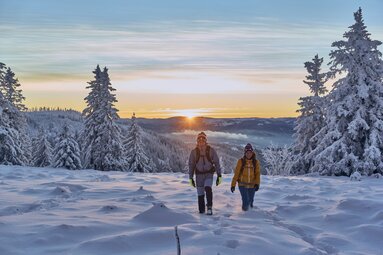 Eine Frau und ein Mann wandern durch den Schnee, der ihnen bis zur Mitte des Schienbeins reicht. Hinter ihnen verschwindet die Sonne hinter einem anderen Gipfel. | © Bodenmais Tourismus & Marketing GmbH