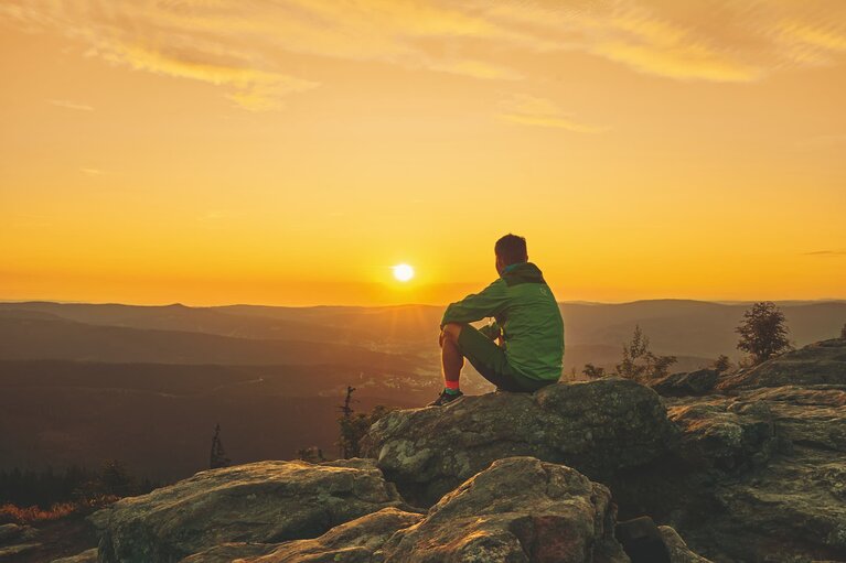 Ein Mann sitzt am Gipfel des großen Arbers und schaut zur aufgehenden Sonne. | © Bodenmais Tourismus & Marketing GmbH