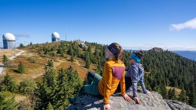 Zwei Wanderer sitzen am Großen Arber und blicke zu den Radartürmen am Gipfel. | © Bodenmais Tourismus & Marketing GmbH