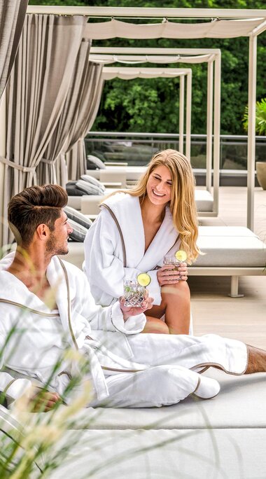 Ein Paar liegt auf einer Terrasse auf einem Day Bett in Bademantel mit einem Cocktail in der Hand | © Günter Standl