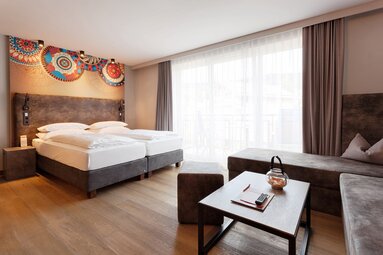 In einem Hotelzimmer steht mittig an der Wand ein Bett mit weißer Bettwäsche, Das Kopfteil ist geschmackvoll mit Mandalas in den Farben orange und blau verziert. Der Rest des Raumes, wie die Sofalandschaft gegenüber vom Bett, ist in neutralen Tönen gehalten. | © Bodenmais Tourismus & Marketing GmbH