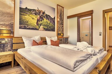 Zu sehen ist ein Hotelzimmer. Ein Bett mit weißer Bettwäsche steht an einer Wand, an der ein Bild von einer Kuh, die auf der Weide steht, hängt. Hinter den Nachtkästchen hängen hohe schmale Bilder aus demWald. | © Bodenmais Tourismus & Marketing GmbH