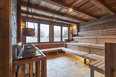 Der Blick fällt in eine Sauna mit Glasfront. Über dem Saunaofen an der Seite hängt ein bronzefarbener Kessel. | © Bodenmais Tourismus & Marketing GmbH