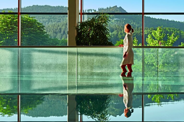 Eine Frau in Bademantel läuft hinter einem spiegelglatten Becken vorbei und blickt durch das große Fenster hinauf auf den Silberberg. | © Bodenmais Tourismus & Marketing GmbH