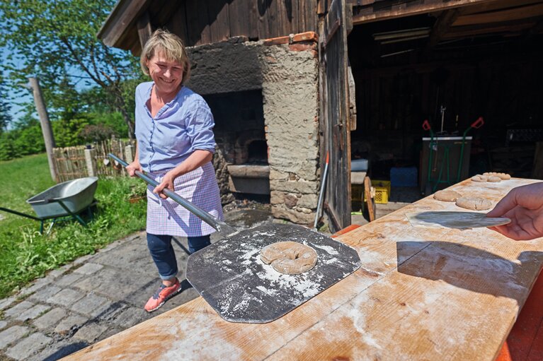 Eine lächelnde Frau hält einen Brot- & Pizzaschieber, auf dem ein rundgeformter Brotteig liegt, in den Händen. Sie befindet sich draußen und hinter ihr steht ein großer Steinofen | © Bodenmais Tourismus & Marketing GmbH