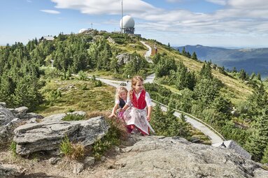 Zwei Mädchen in Dirndl wandern auf den Großen Arber. Hinter ihnen stehen die Radartürme und die Sonne scheint. | © Bodenmais Tourismus & Marketing GmbH