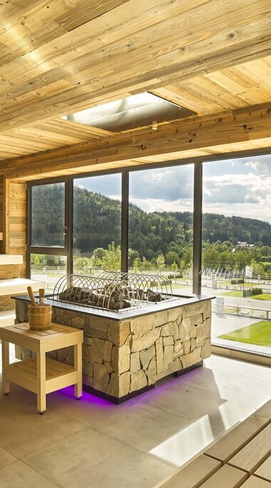 Der Blick fällt in eine Saune, die komplett mit hellem Holz verkleidet ist. In der Mitte steht der Saunaofen. Dahinter kann man durch eine große Fensterfront auf die Parzellen des Camping Resort schauen. | © Bodenmais Tourismus & Marketing GmbH