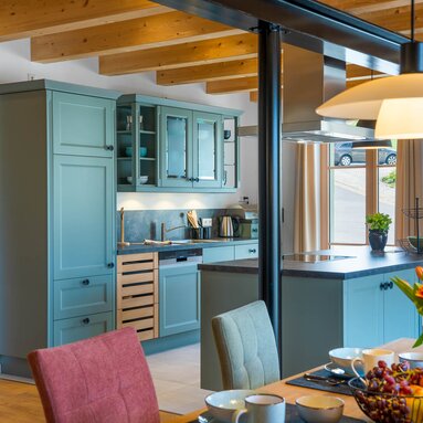 Gemütlicher Wohnraum einer Ferienwohnung mit heller petrol-farbener Küche im Landhausstil, sowie Holzesstisch mit einer Vase voll Tulpen. | © Bodenmais Tourismus & Marketing GmbH