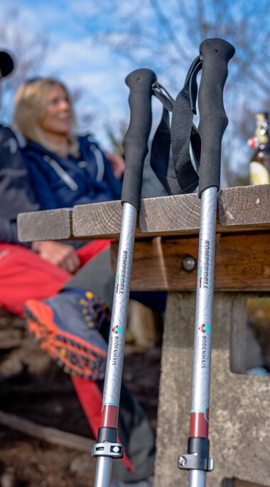 Ein Paar Wanderstöcke im Bodenmais Design ist an einen Picknicktisch gelehnt. Im Hintergrund sitz ein paar an diesem Tisch. | © Bodenmais Tourismus & Marketing GmbH