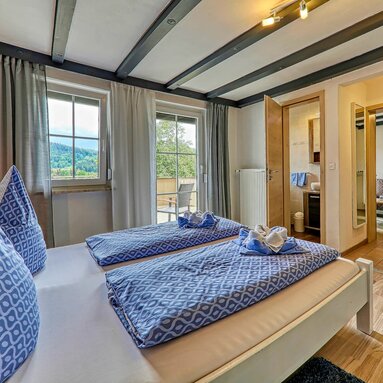 Der Blick fällt über ein mit weiß-blauer Bettwäsche bezogenes Bett. Gegenüber vom Bad steht die Tür zum Badezimmer offen.  | © Bodenmais Tourismus & Marketing GmbH
