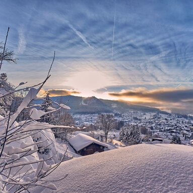 Über glitzernden Schnee hinweg blickt man auf Bodenmais. Über der Ortschaft wacht der Silberberg, hinter dem die Sonne hervor blitzt, der blaue Himmel ist durchzogen von Schleierwolken. | © Bodenmais Tourismus & Marketing GmbH