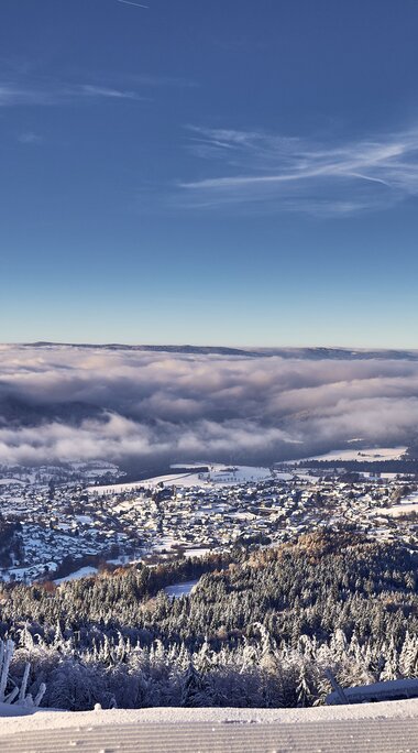 Von einer Loipe aus blickt man herab auf das verschneite Bodenmais, das im Sonnenlicht erstrahlt. Hinter Bodenmais liegt Nebel im Tal. | © Bodenmais Tourismus & Marketing GmbH