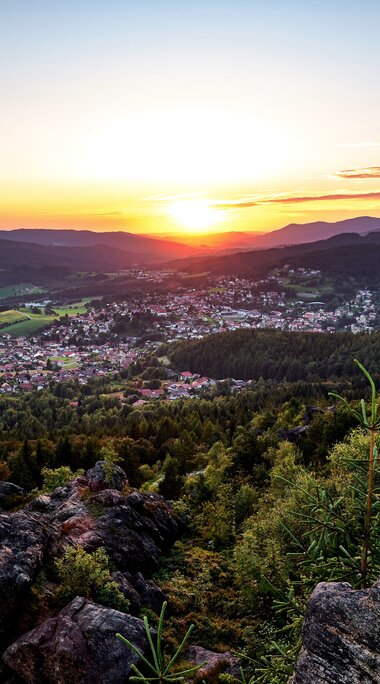 Von einer Anhöhe sieht man die orange Sonne über Bodenmais untergehen. Dunkelgrüne Wälder umgeben die Ortschaft und die umliegenden Berge ergeben einen straken Kontrast zum roten Himmel. | © Bodenmais Tourismus & Marketing GmbH