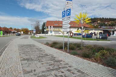 Im Vordergrund ist ein Gehweg mit Beschilderung der Ortschaft. Im Hintergrund sieht man die Ortschaft mit Häusern und einem Parkplatz.  | © Markt Bodenmais