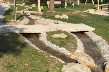 Neben einem Spielplatz verläuft ein künstlich angelegter Bachlauf mit Brücke. Die Brücke besteht aus einer großen Granitplatte. Es sind große Steine zwischen dem Wasser.  | © Markt Bodenmais