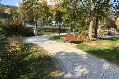 Ein aus Granitsteinen gepflasterter Weg verläuft in einer Parkanlage mit Bäumen, Grünfächen und Bänken. Im Hintergrund sieht man den Zaun der Tennisanlage.  | © Markt Bodenmais