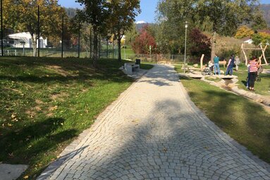 Ein aus Granitsteinen gepflasterter Weg verläuft in einer Parkanlage mit Bäumen, Grünfächen und Bänken. Links liegt der Tennisplatz, rechts der Spielplatz und im Hintergrund sieht man das Freibad.  | © Markt Bodenmais