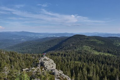 Von weiter weg sieht man ein Paar auf dem Großen Arber stehen, welches den Blick über die Arberregion schweifen lässt. | © Bodenmais Tourismus & Marketing GmbH