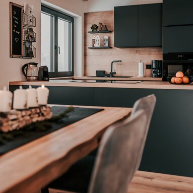 Über den Esstisch hinweg blickt man auf die moderne, dunkle Küche. | © Bodenmais Tourismus & Marketing GmbH