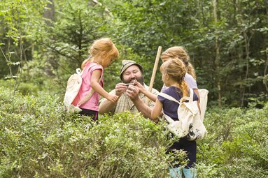 Ein lachender Mann mit Vollbart, grünem Filzhut und kariertem Hemd (auch Woid Woife genannt) zeigt drei Mädchen ein Stück Holz, das er in den Händen hält. Die vier Personen befinden sich in einem Wald. Die Kinder tragen einen Jute Rucksack auf dem Rücken, auf dem der Woid Woife gedruckt ist. | © Bodenmais Tourismus & Marketing GmbH