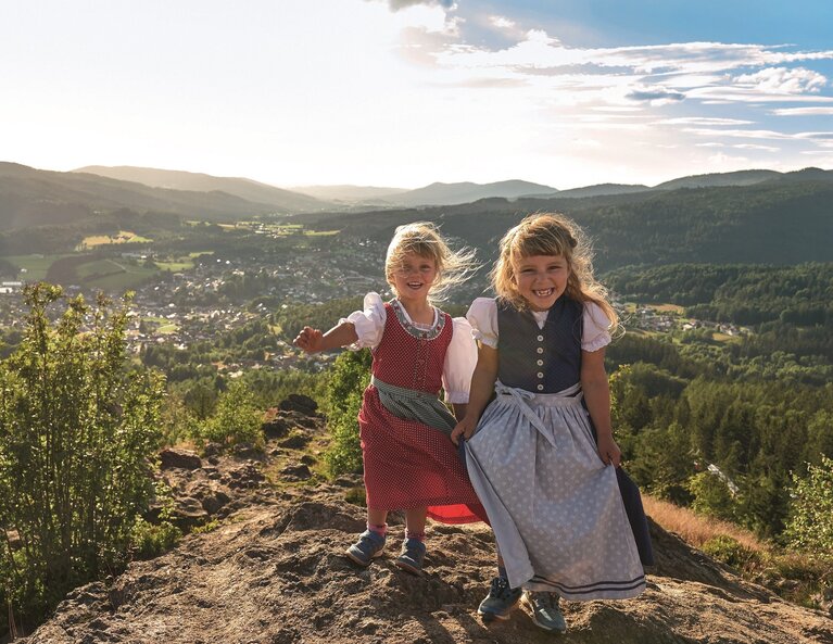 Zwei Mädchen stehen lachend im Dirndl auf einem Berg und die langsam untergehende Sonne strahlt sie von hinten an. Im Tal liegt Bodenmais. | © Bodenmais Tourismus & Marketing GmbH