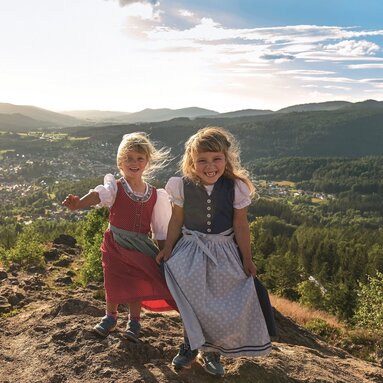 Zwei Mädchen stehen lachend im Dirndl auf einem Berg und die langsam untergehende Sonne strahlt sie von hinten an. Im Tal liegt Bodenmais. | © Bodenmais Tourismus & Marketing GmbH