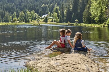 Drei Mädchen sitzen auf einem Stein der flach aus dem Wasser ragt. Sie tragen Dirndl und halten ihre Füße in den See. Am gegenüberliegenden Ufer steh eine Hütte, dahinter liegt Wald. | © Bodenmais Tourismus & Marketing GmbH