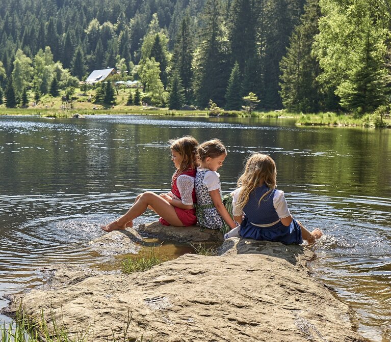 Drei Mädchen sitzen auf einem Stein der flach aus dem Wasser ragt. Sie tragen Dirndl und halten ihre Füße in den See. Am gegenüberliegenden Ufer steh eine Hütte, dahinter liegt Wald. | © Bodenmais Tourismus & Marketing GmbH