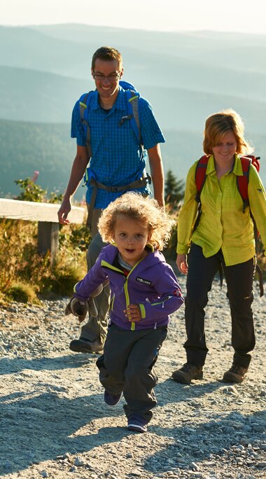 Eine glücklich aussehende Familie bestehend aus Vater, Mutter und zwei kleinen Kindern wandert auf einem Schotterweg, der von zwei Holzbalken von den Wiesen getrennt liegt. Im Hintergrund liegt das Tal und weitere Gipfel des Bayerischen Waldes. | © Bodenmais Tourismus & Marketing GmbH