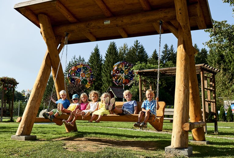 7 Kinder sitzen auf einer großen, hölzernen Hollywood Schaukel. Einige Kinder strecken die Füße, wie beim Schaukeln, nach vorne. Im Hintergrund stehen runde Glasskulpturen | © Bodenmais Tourismus & Marketing GmbH