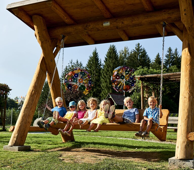 7 Kinder sitzen auf einer großen, hölzernen Hollywood Schaukel. Einige Kinder strecken die Füße, wie beim Schaukeln, nach vorne. Im Hintergrund stehen runde Glasskulpturen | © Bodenmais Tourismus & Marketing GmbH
