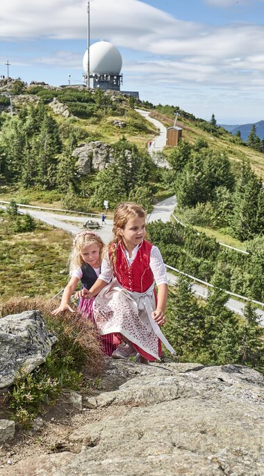 Zwei Mädchen in Dirndl wandern auf den Großen Arber. Hinter ihnen stehen die Radartürme und die Sonne scheint. | © Bodenmais Tourismus & Marketing GmbH