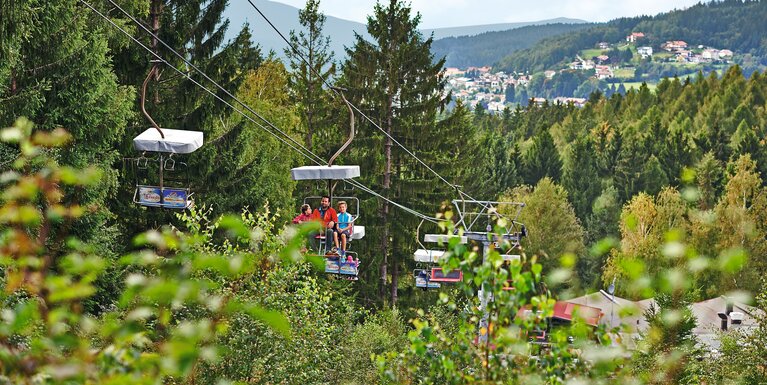 Eine Familie fährt in einem Sessellift auf den Silberberg. Der Blickwinkel ist durch Bäume und Äste hindurch. Im Hintergrund liegt Bodenmais | © Bodenmais Tourismus & Marketing GmbH