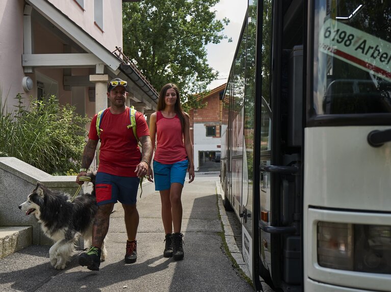 Eine Frau und ein Mann in Wanderklamotten laufen mit einem Husky zu der offenstehenden Tür eines Linienbusses. An dessen Frontscheibe steht: 6198 Arber Bergbahn | © Bodenmais Tourismus & Marketing GmbH