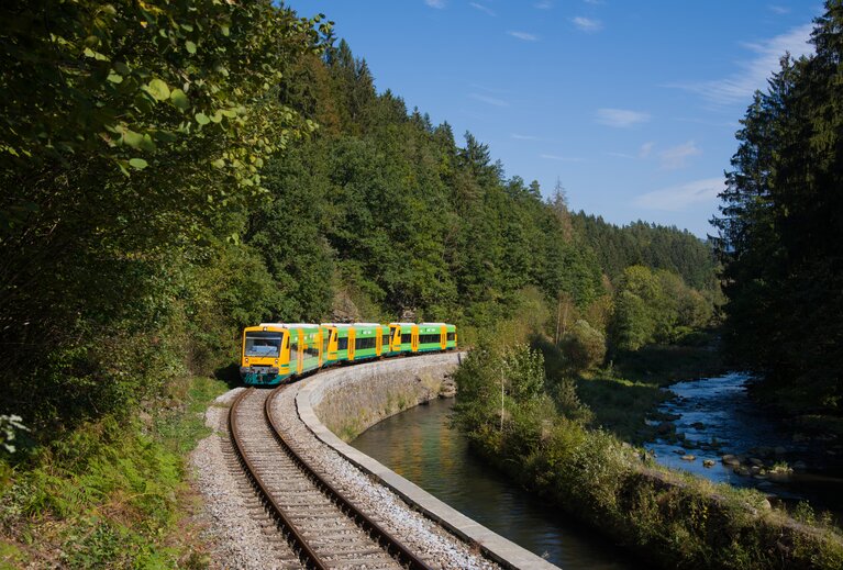 Ein gelb-grüner Zug fährt zwischen einem Waldstück und einem Fluss. | © Bodenmais Tourismus & Marketing GmbH