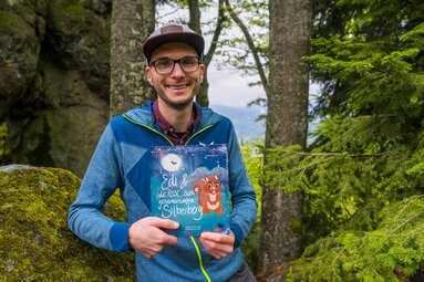 Ein Mann mit Brille und Cap lacht und hält das Kinderbuch "Edi und die Reise zum geheimnisvollen Silberberg in den Händen. | © Bodenmais Tourismus & Marketing GmbH