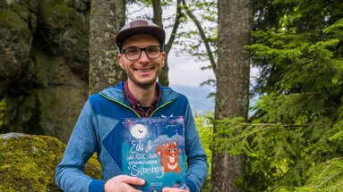 Ein Mann mit Brille und Cap lacht und hält das Kinderbuch "Edi und die Reise zum geheimnisvollen Silberberg in den Händen. | © Bodenmais Tourismus & Marketing GmbH