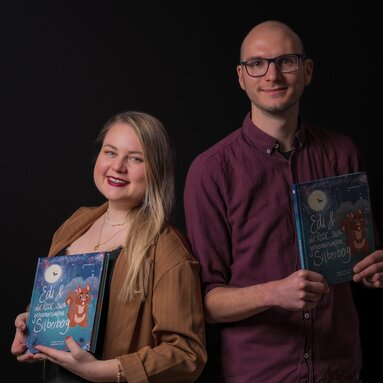 Eine blonde Frau und ein Mann mit Brille, Grafikerin und Autor des Bodenmaiser Kinderbuchs, stehen mit besagtem Buch vor einer schwarzen Wand | © Bodenmais Tourismus & Marketing GmbH