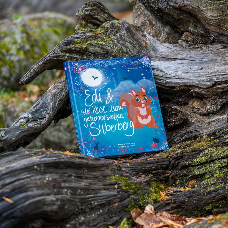Das Bodenmaiser Kinderbuch "Edi und die Reise zum geheimnisvolle Reise zum Silberberg" steht auf einem mit moosbewachsenem,  alten Baumstamm. | © Bodenmais Tourismus & Marketing GmbH