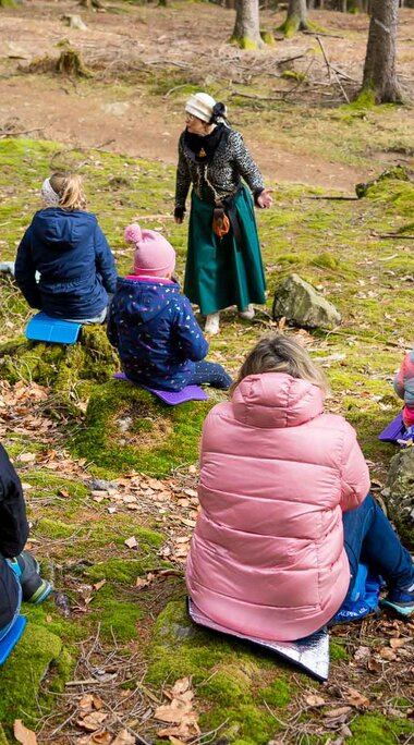 Mehrere Kinder und ein paar Elternteile sitzen auf Baumstümpfen in einem moosigem Waldabschnitt. Sie schauen alle auf eine Frau, die eine märchenhafte Verkleidung trägt und ihnen eine Geschichte erzählt. | © Bodenmais Tourismus & Marketing GmbH