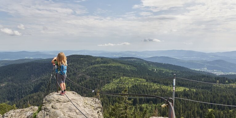 Ein Mädchen steht auf einem Gipfel, der mit zwei Stahlseilen zum Abgrund gesichert ist, und blickt in die Ferne auf den Bayerischen Wald. | © Bodenmais Tourismus & Marketing GmbH